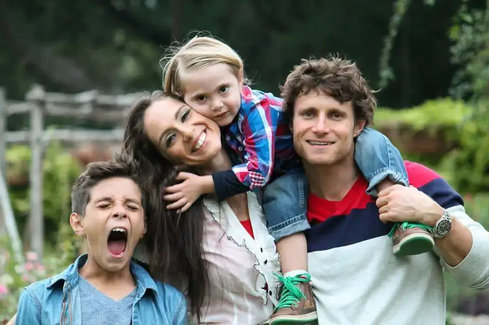 A family stands together in a garden, smiling for a photo, surrounded by flowers and greenery.